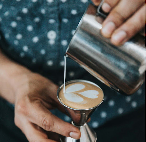 A barista's hands carefully pouring steamed milk into a ceramic cup to create latte art, in a warm, dimly lit cafe